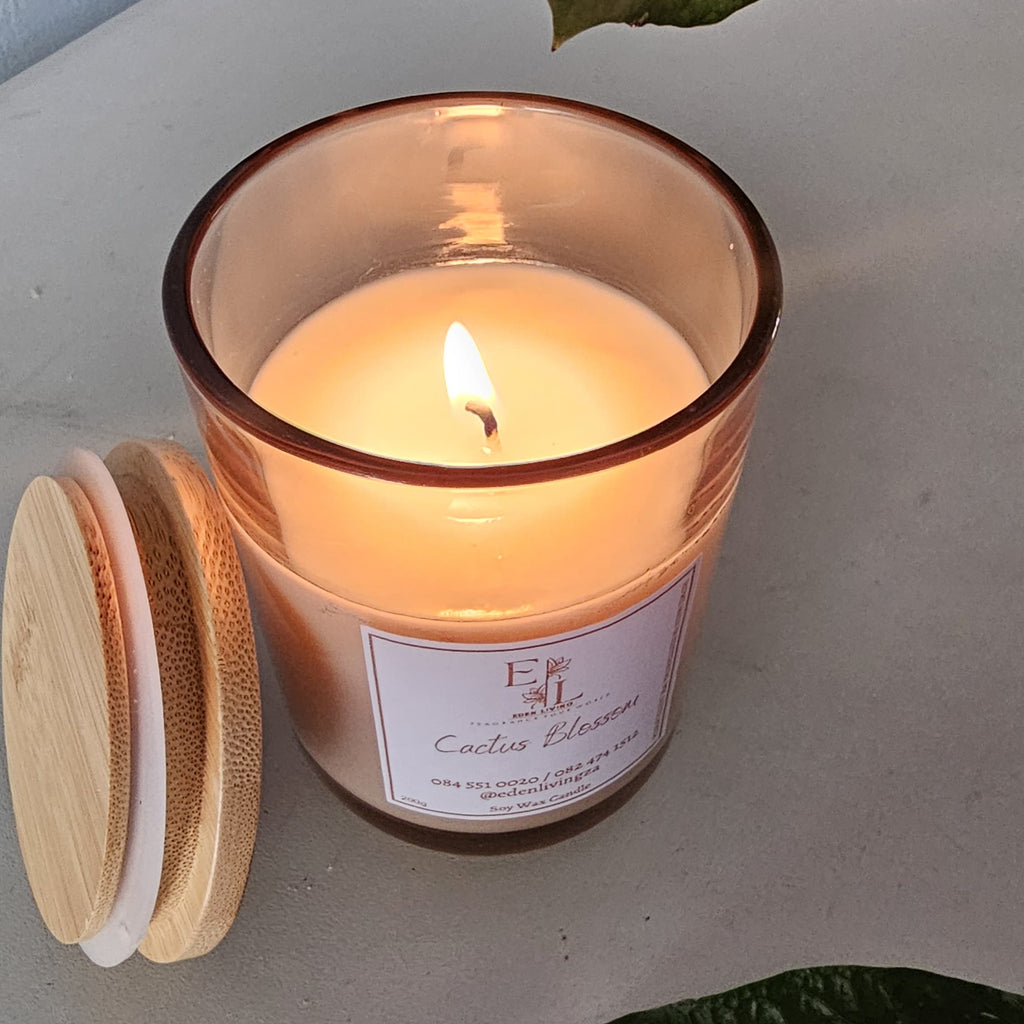 Lit candle in a glass jar with wooden lid on the side, surrounded by leaves.